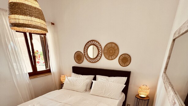 Een opgemaakt bed met witte lakens en opgerolde handdoeken, een geweven hanglamp en decoratieve spiegelwand.