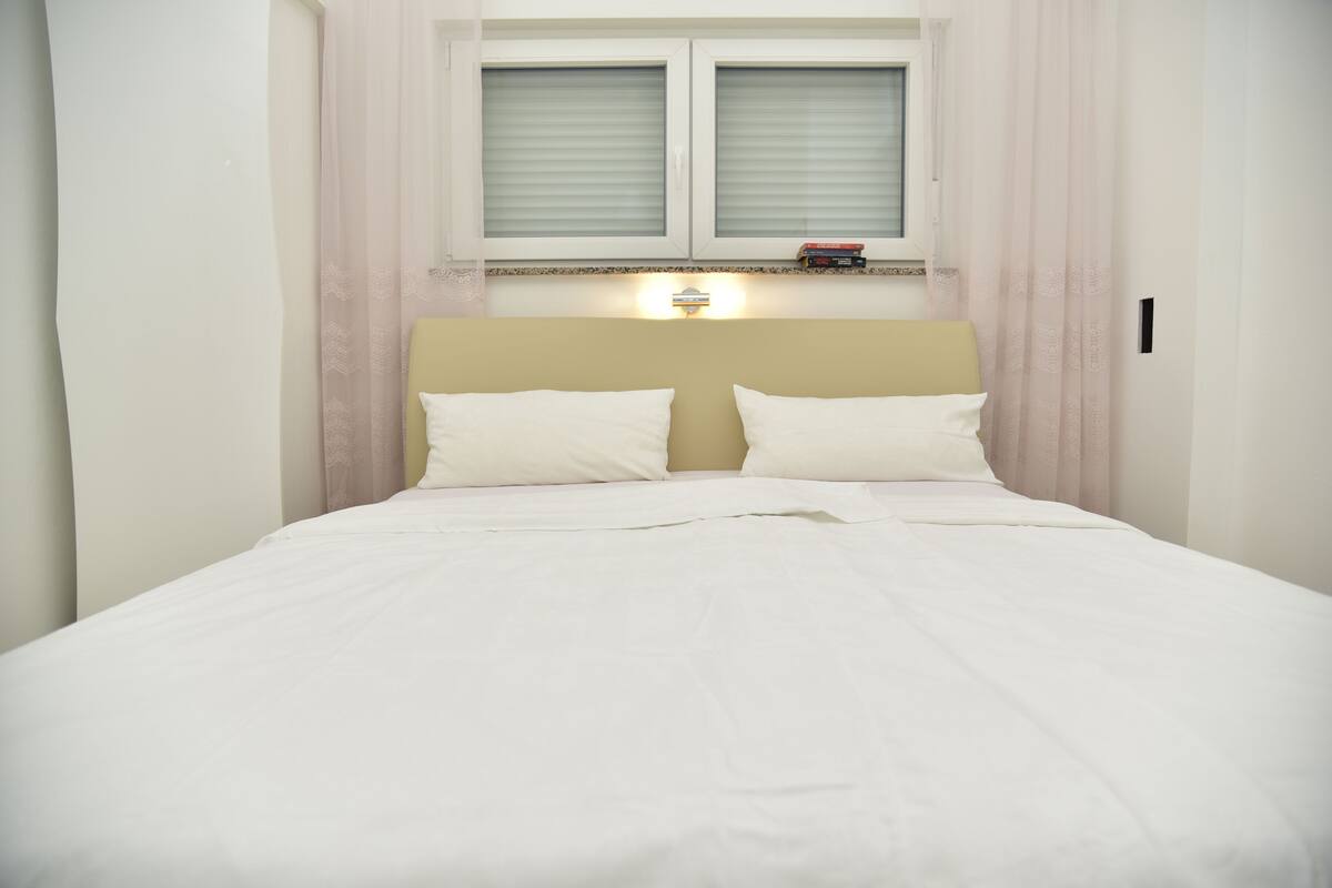 Un lit bien fait, avec du linge de lit blanc, une tête de lit beige et une fenêtre équipée de stores.