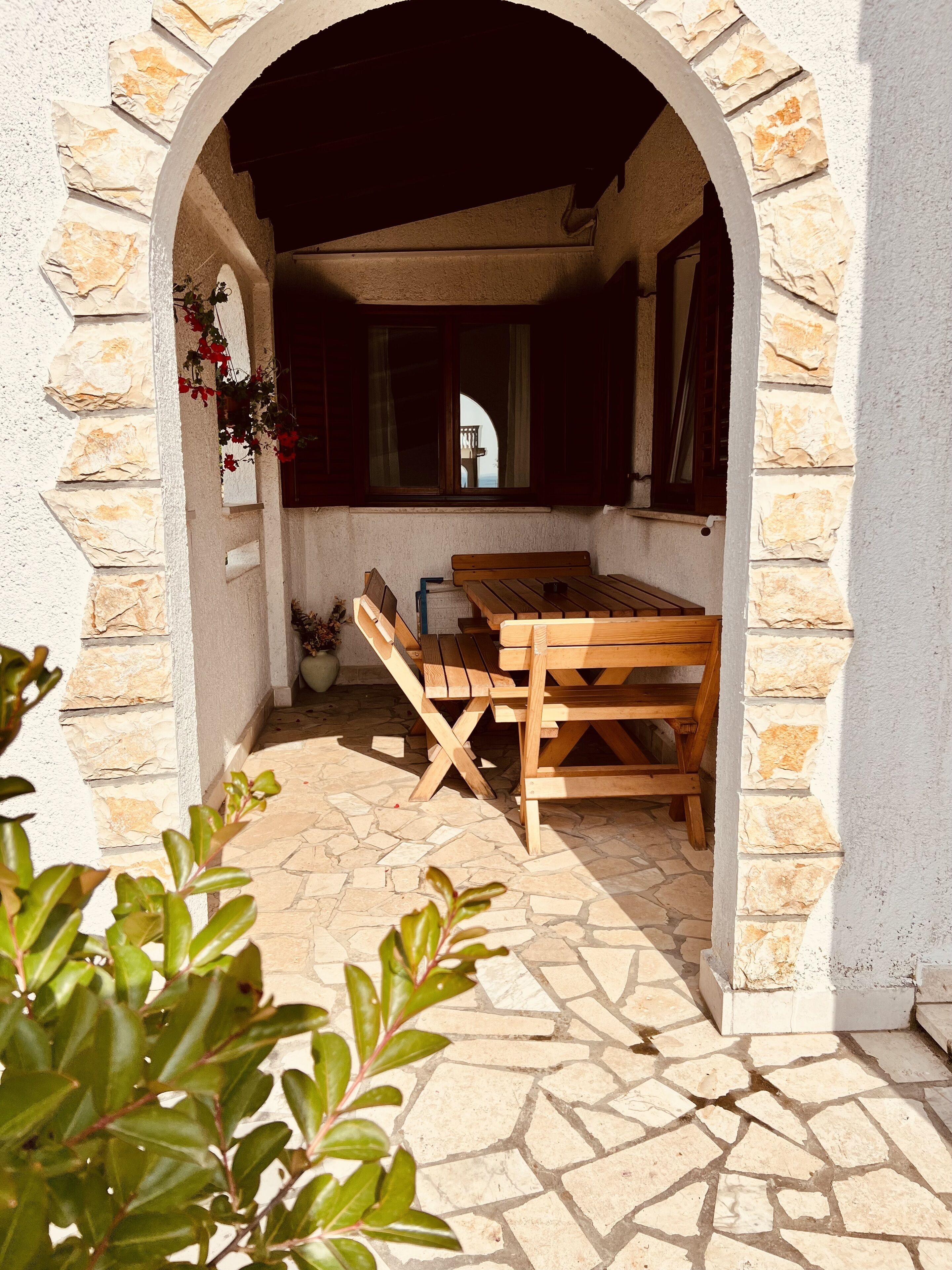 An outdoor seating area with a wooden table and benches, stone walls, and a view through an archway.