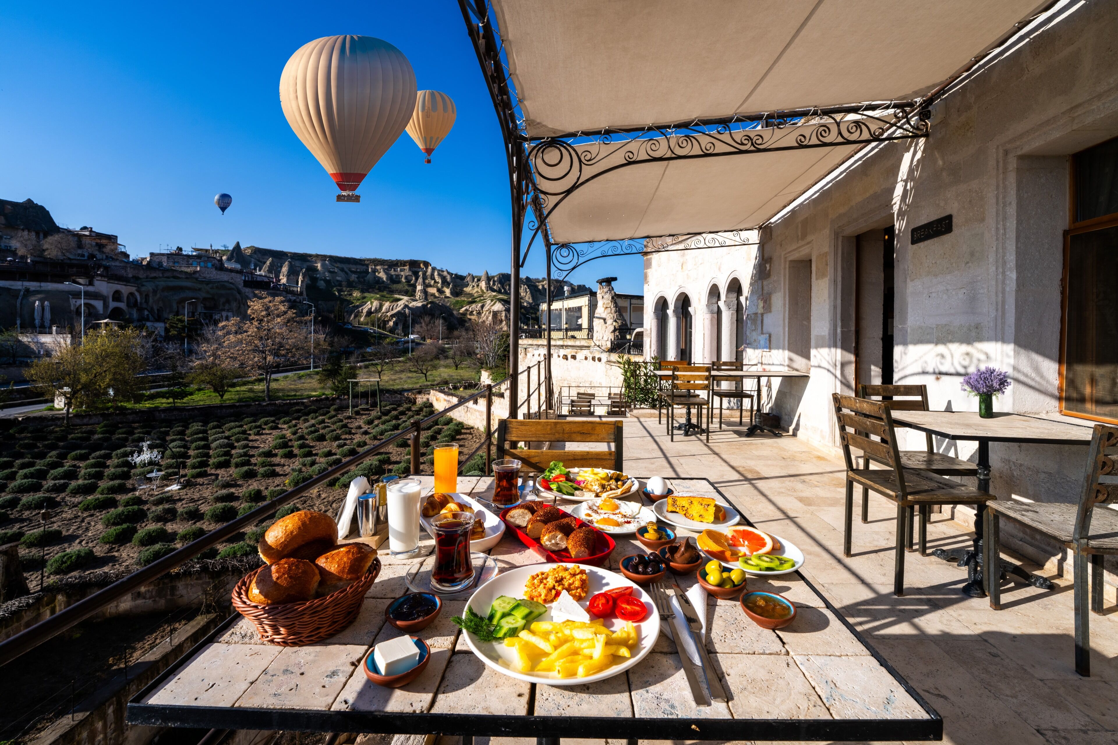 A table set with a variety of foods and drinks, overlooking a scenic landscape with buildings and hot air balloons.