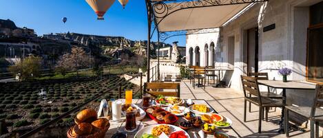 A table set with a variety of foods and drinks, overlooking a scenic landscape with buildings and hot air balloons.