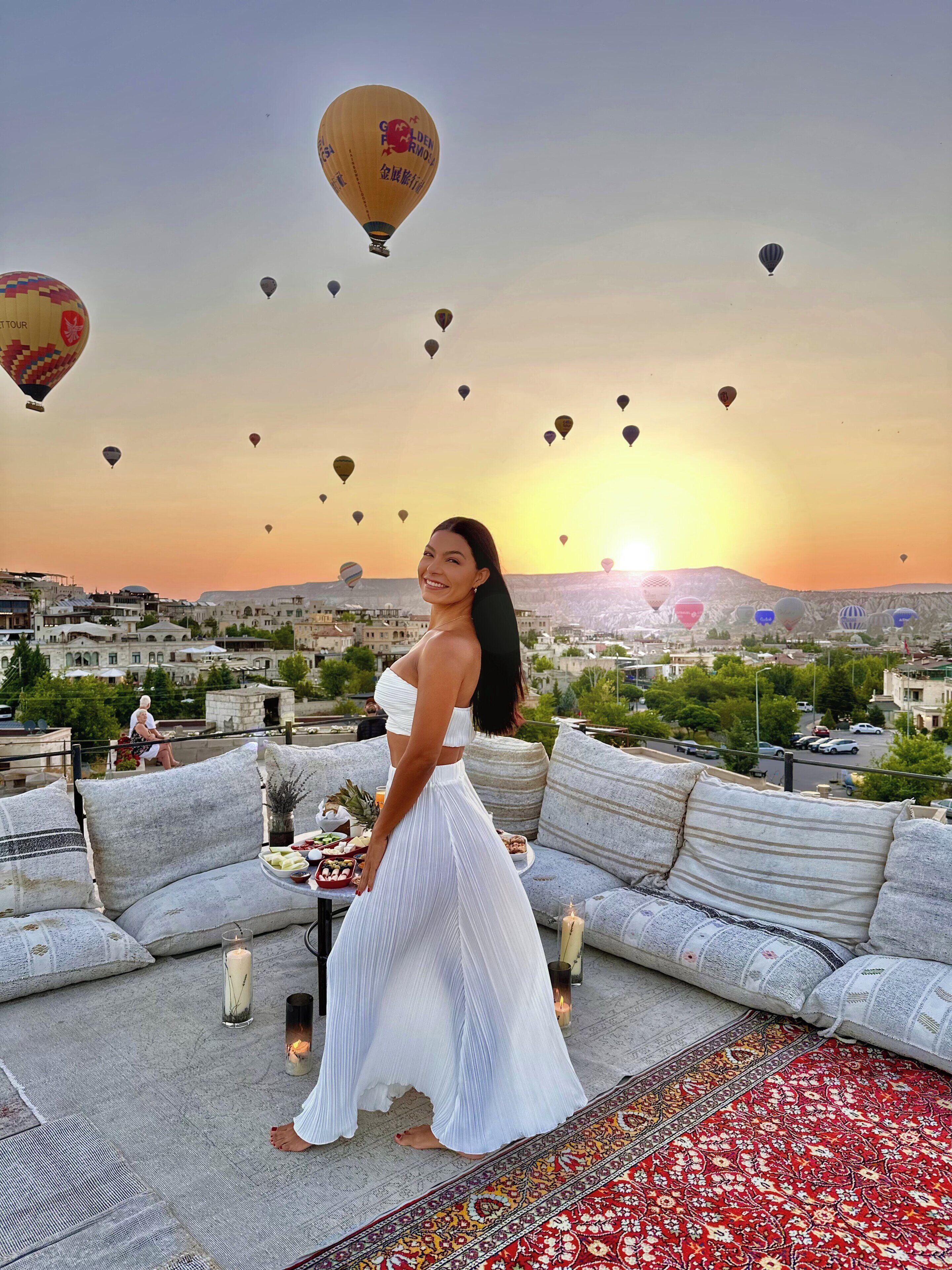 A woman standing on a rooftop terrace with a cityscape and hot air balloons in the background.