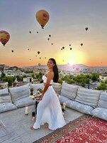 Una mujer parada en una terraza en la azotea, con un paisaje urbano y globos aerostáticos al fondo.