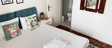 Una cama bien hecha con ropa de cama blanca, un cabecero color turquesa y almohadones decorativos. Se ve una mesita de noche con una planta y una cesta. La habitación tiene piso de madera y un espejo en la pared.
