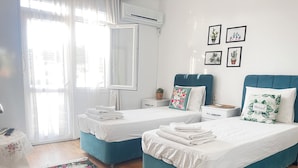 A hotel room with two beds, a large window with sheer curtains, a floral rug, and a wall with framed pictures. - Siren Pansiyon (Foça)