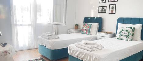 A hotel room with two beds, a large window with sheer curtains, a floral rug, and a wall with framed pictures.