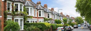 A row of traditional brick houses with white trim and pitched roofs, parked cars along the street, and lush greenery. - RED F Large Studio (London)