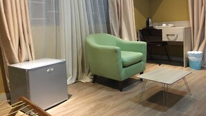 Habitación pequeña con un sillón verde, un escritorio con una silla, un televisor de pantalla plana en la pared y un refrigerador compacto.