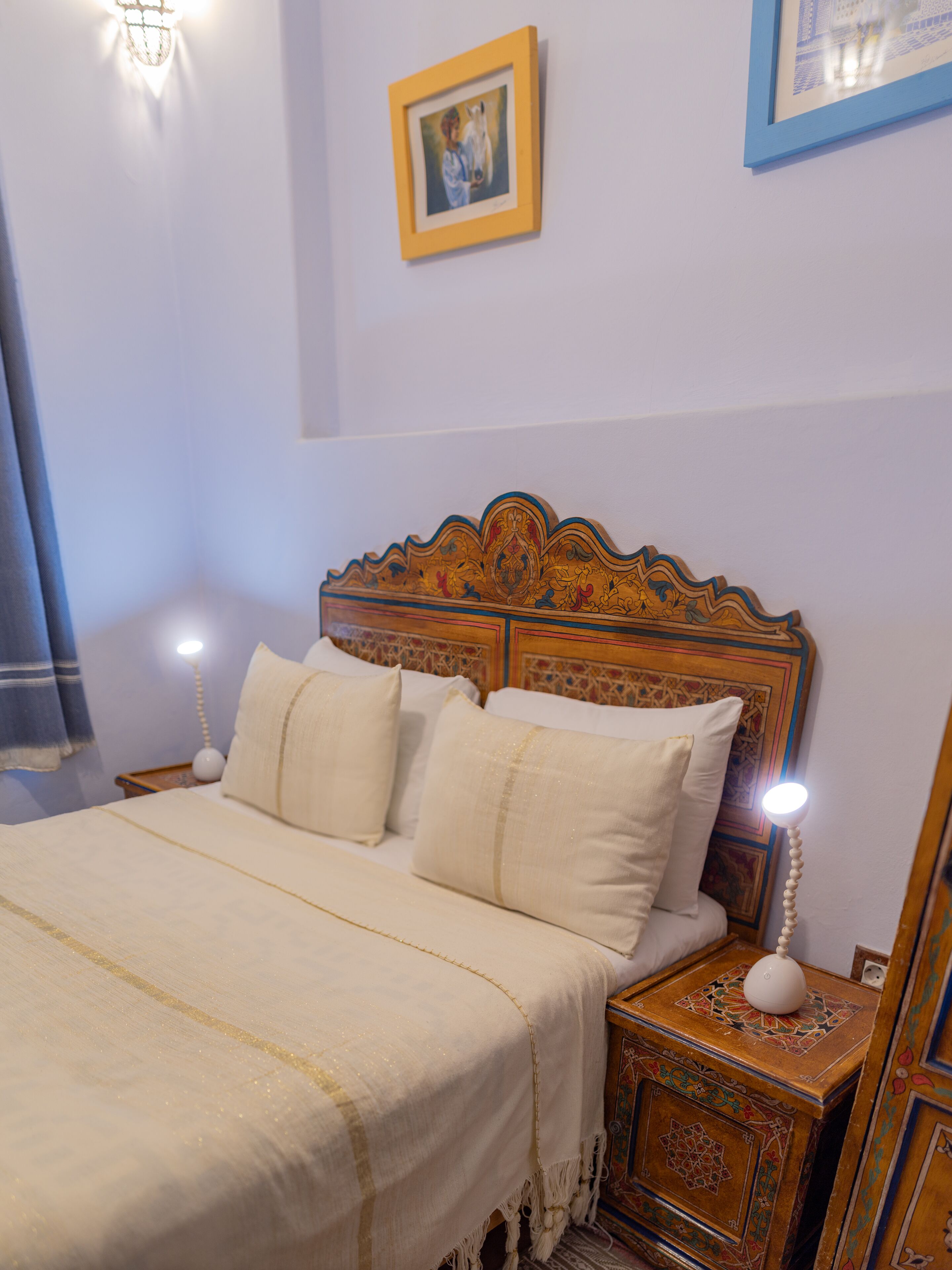 A neatly made bed with a white bedspread and pillows, a wooden headboard with intricate carvings, and a small bedside table with a lamp.