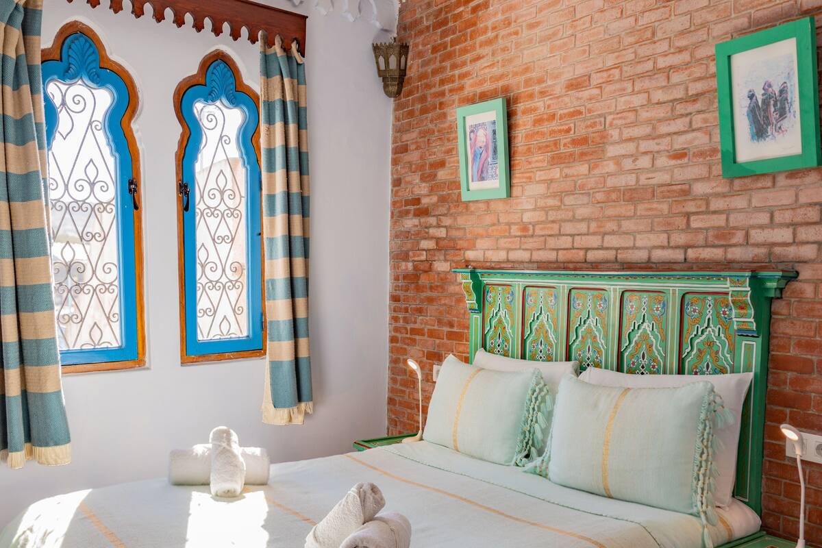 Une pièce avec un mur en briques, un lit doté d’une tête de lit verte et deux fenêtres ornées de rideaux décoratifs.