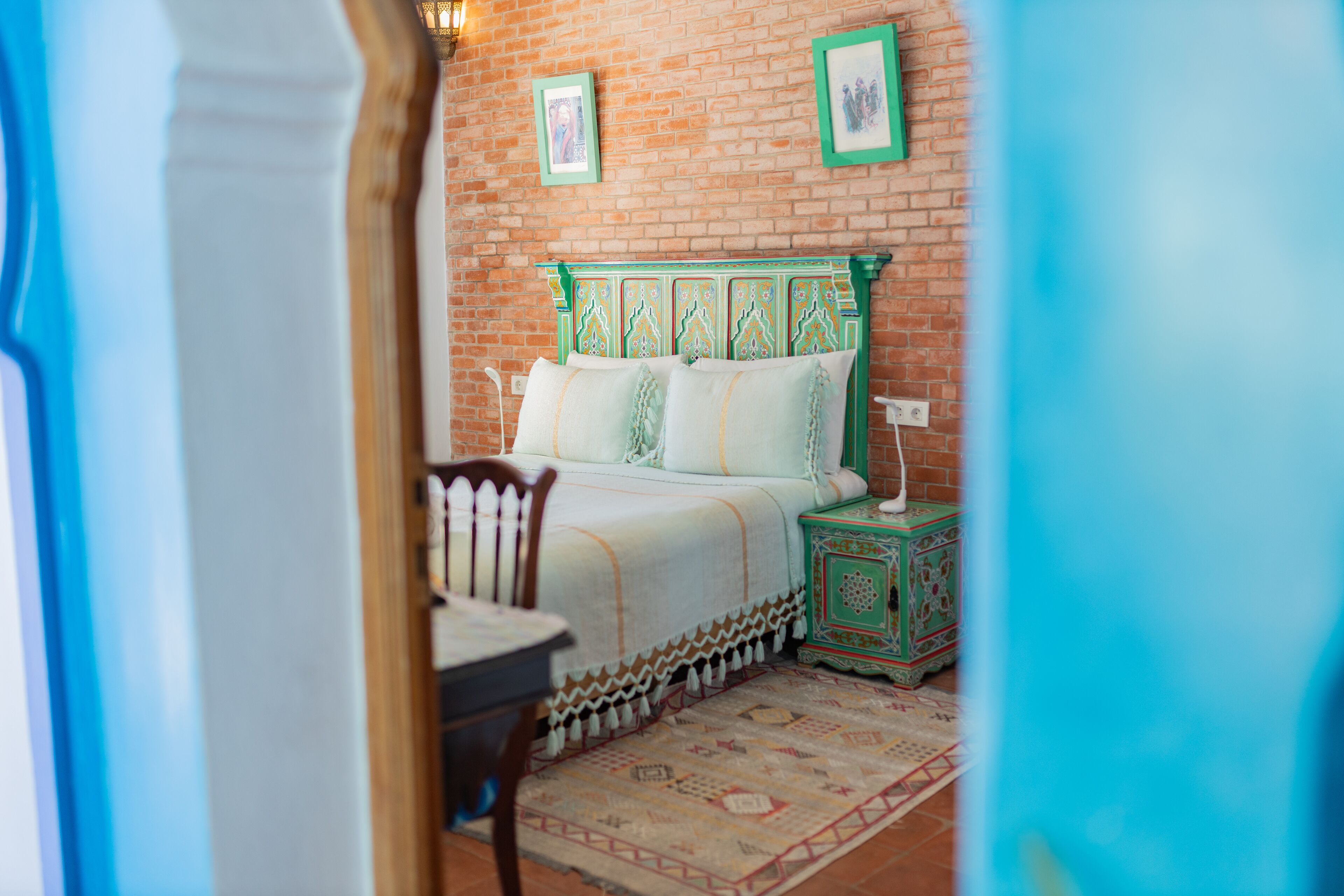 Una habitación con pared de ladrillo, cama con cabecera verde, una mesita y una silla.