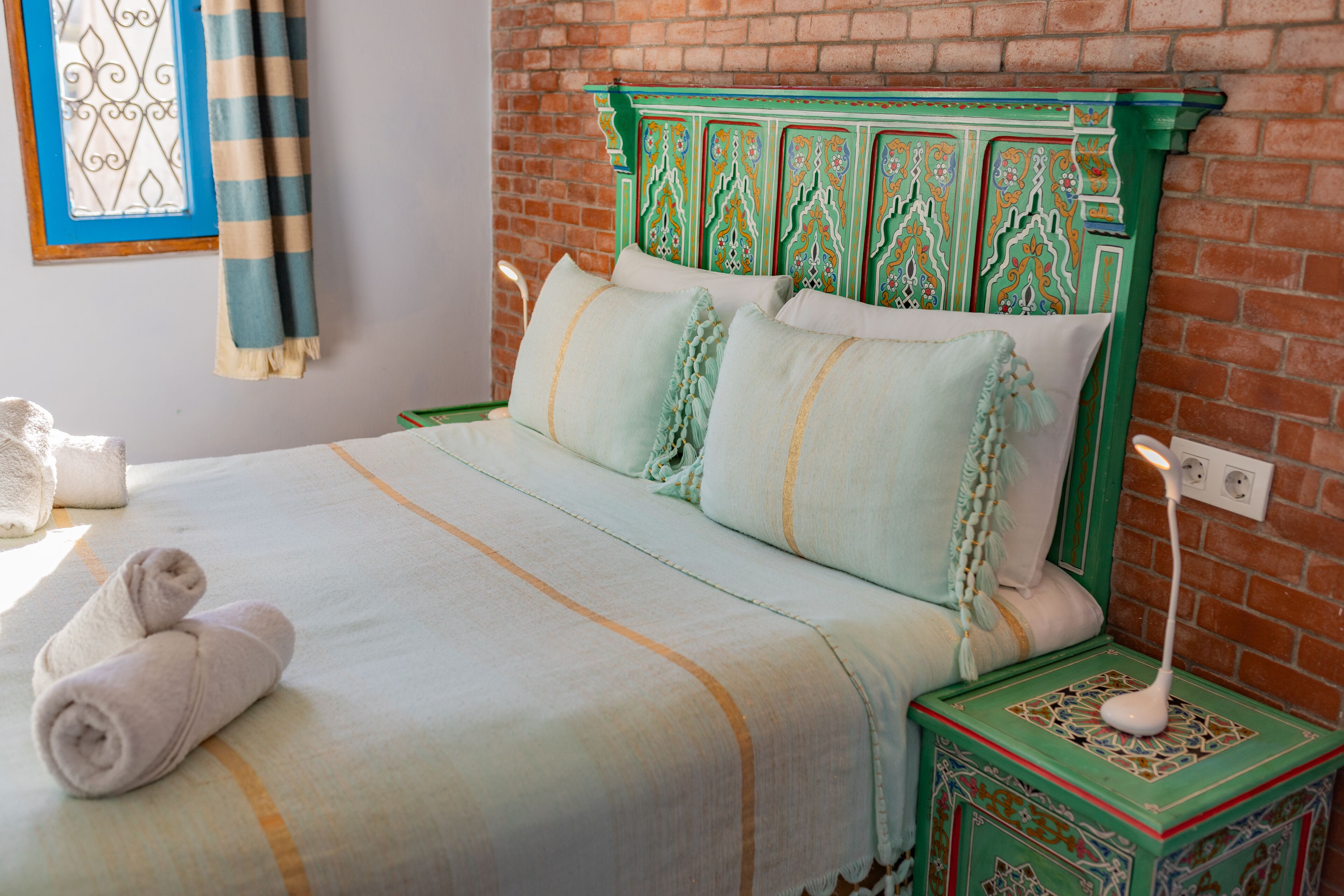 A neatly made bed with a striped blanket and pillows, a bedside table with a lamp, and a decorative headboard against a brick wall.