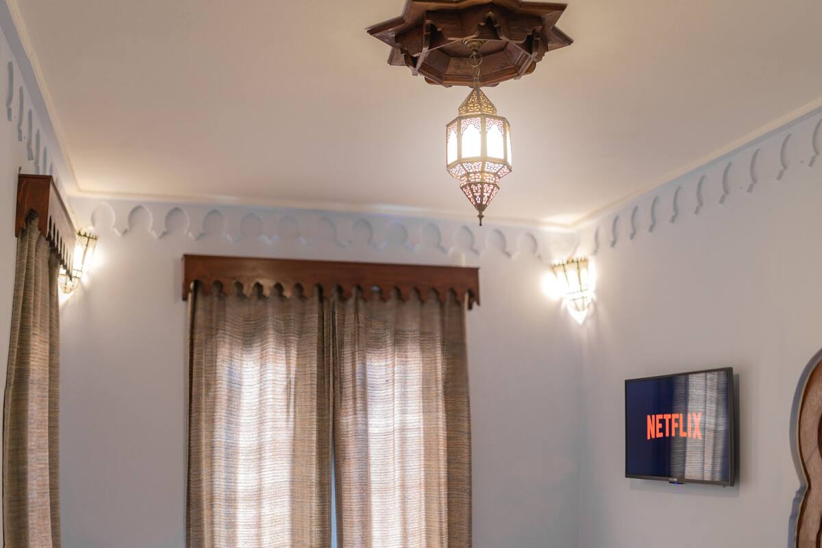 Une chambre à coucher avec un lit, des rideaux, un téléviseur fixé au mur et un luminaire décoratif au plafond.