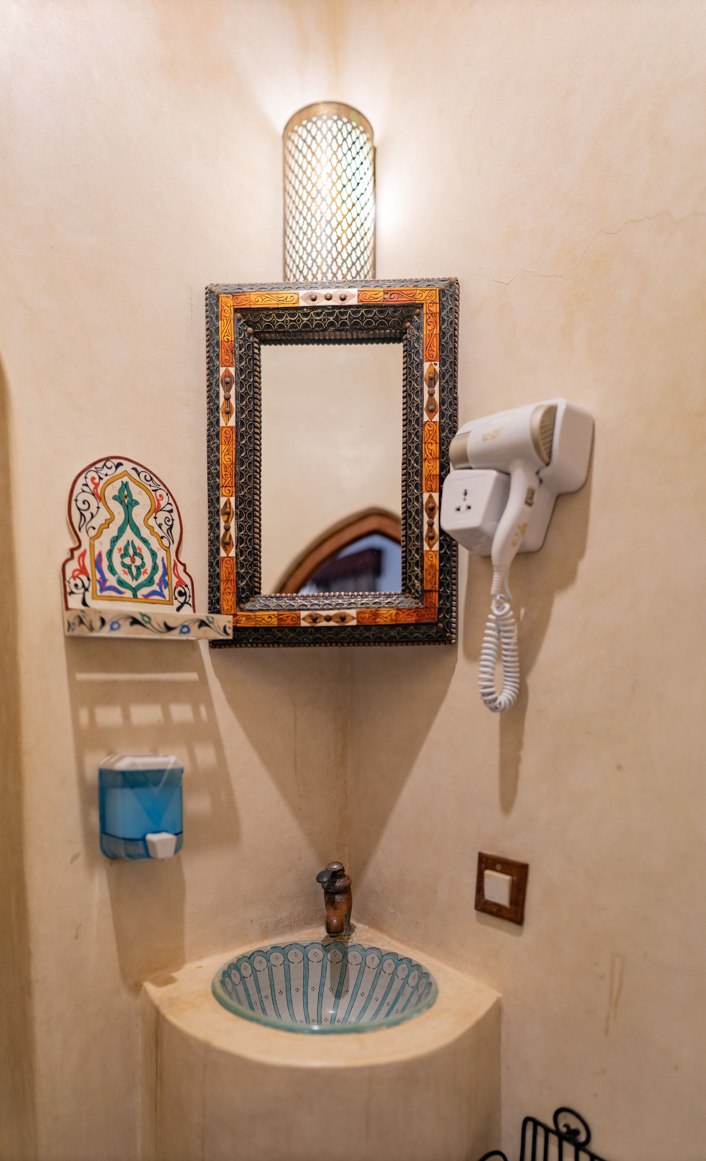 A bathroom with a decorative mirror, a hairdryer, a sink with a blue basin, and a soap dispenser.