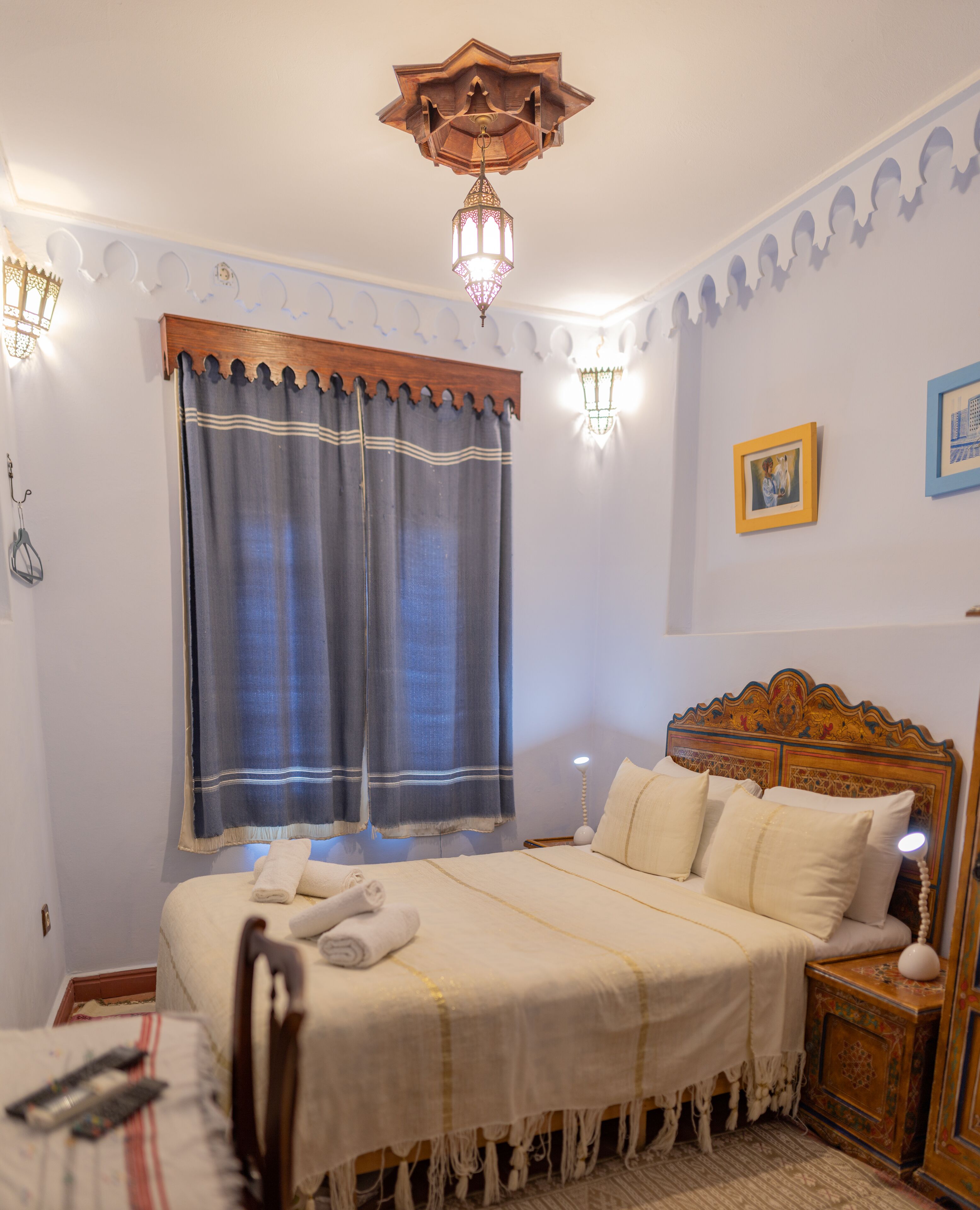 Une chambre à coucher bien rangée, avec un grand lit, une tête de lit en bois, une table de chevet, une lampe fixée au mur et une fenêtre avec des rideaux.