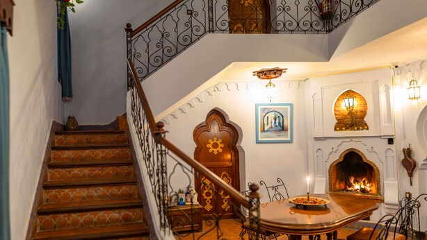 Un escalier orné de balustrades travaillées, un coin repas avec une table et des chaises, et une cheminée avec un manteau en briques.