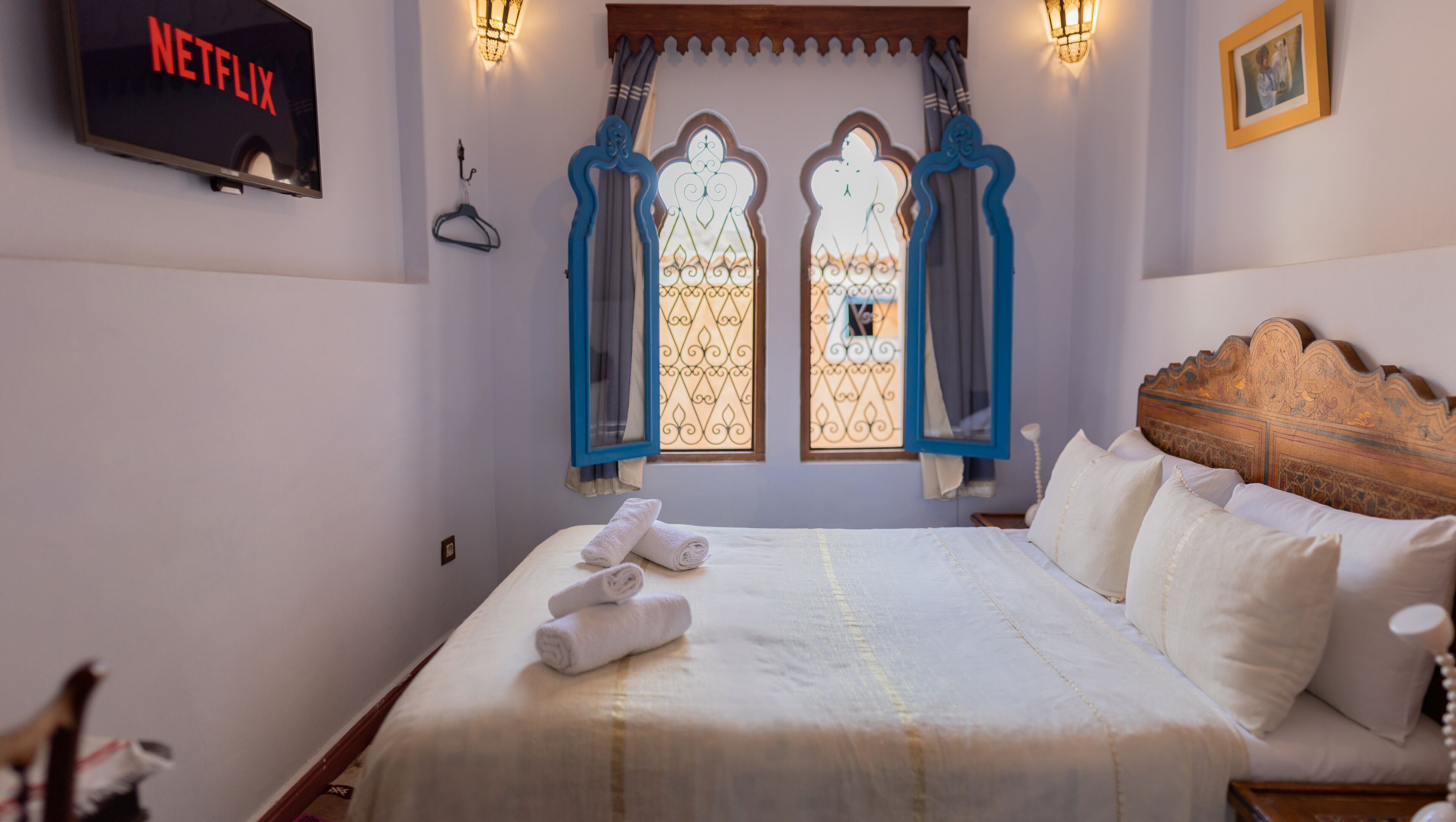 A neatly made bed with white linens and rolled towels, a wall-mounted TV displaying the Netflix logo, and a window with decorative blue curtains.