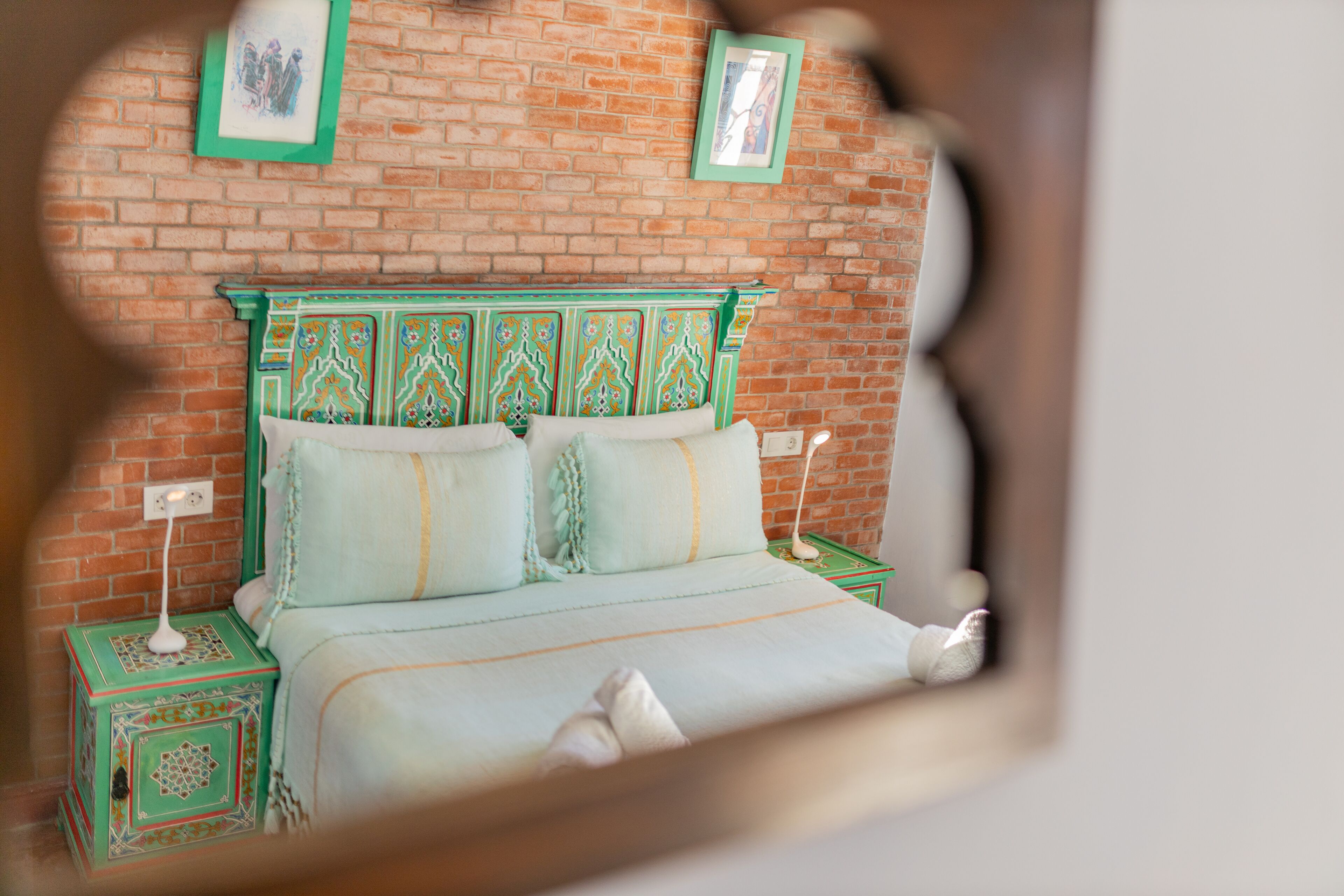 Una cama con cabecera verde y ropa de cama blanca, una mesita de noche con lámpara, y una pared de ladrillo con cuadros enmarcados.