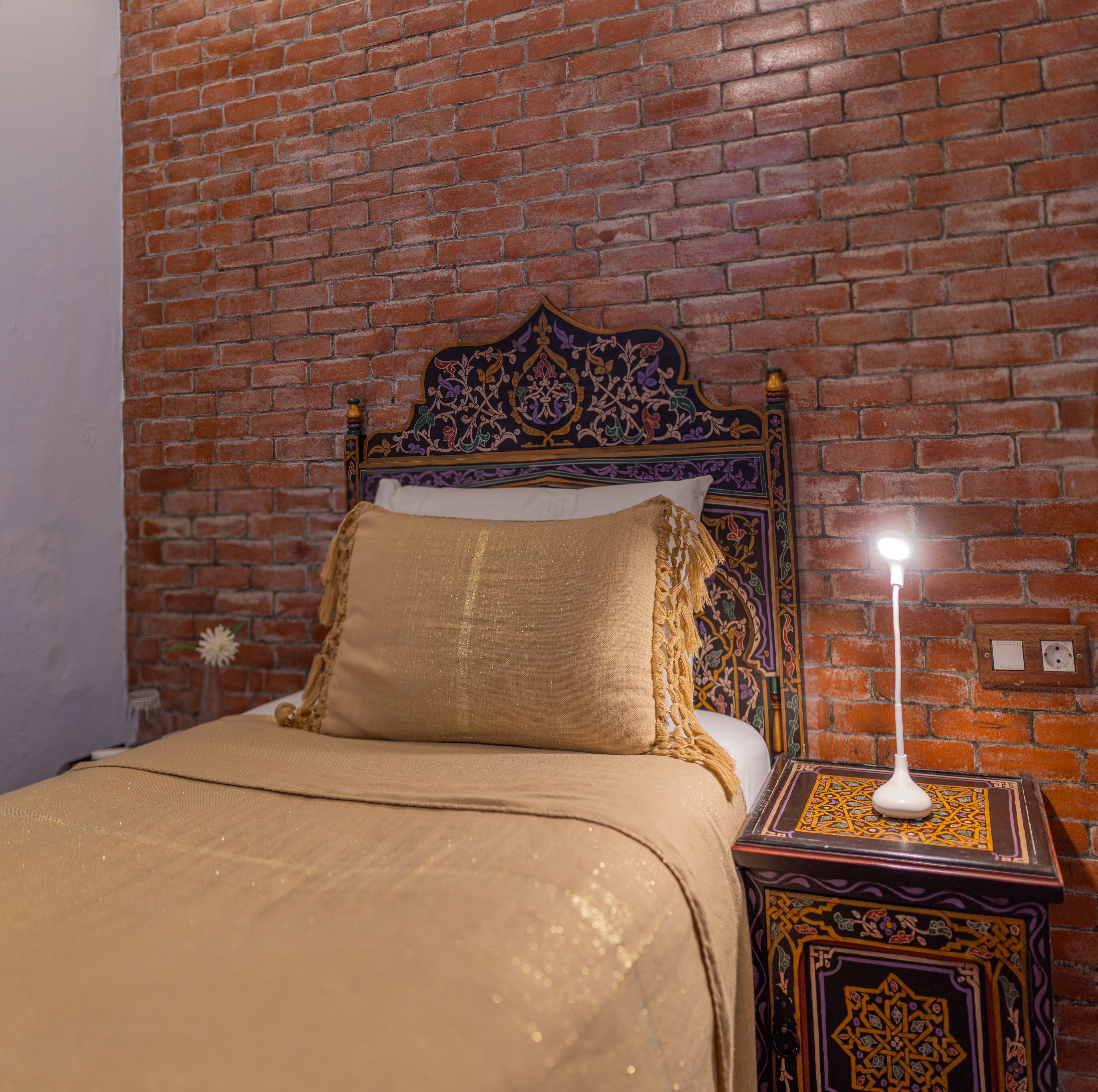 A bed with a headboard against a brick wall, a bedside table with a lamp, and a decorative wall plate.