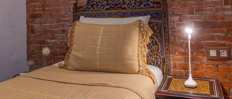 A bed with a headboard against a brick wall, a bedside table with a lamp, and a decorative wall plate.