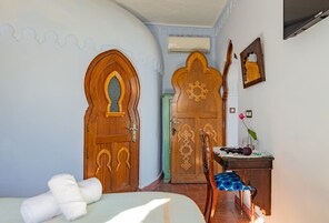 Una habitación con dos puertas de madera, una cama con cubrecama blanco, un escritorio con un florero y una silla con cojín azul.