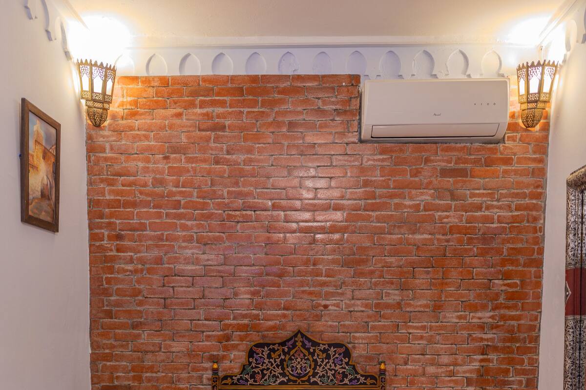 Une chambre d’hôtel avec un mur en briques, un lit avec une tête de lit, une table de chevet, une lampe et un climatiseur fixé au mur.