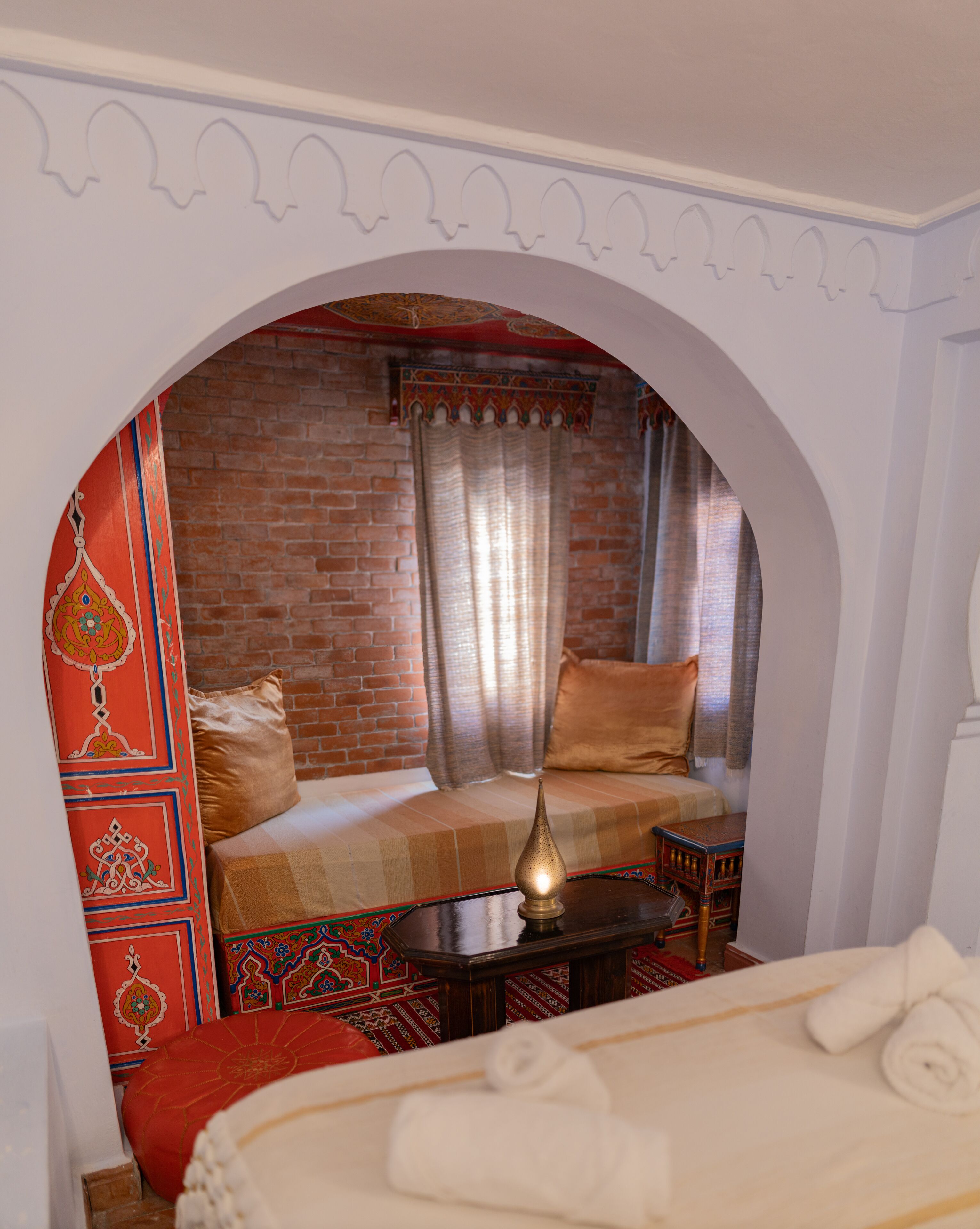 A room with a brick wall, a bed with a red and beige patterned bedspread, a small table with a lamp, and a decorative pillow.