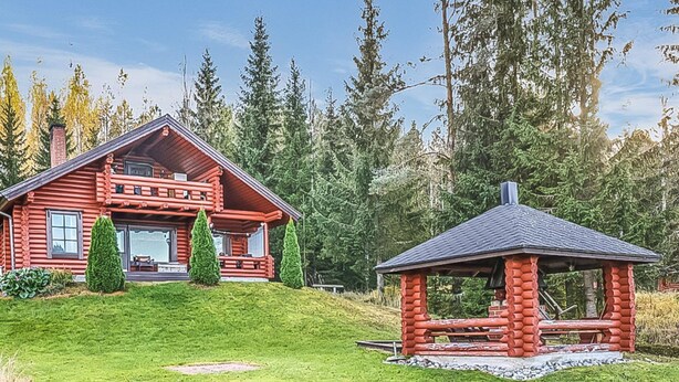 A log cabin with a porch, a small gazebo, and a grassy yard surrounded by trees.