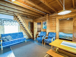 Een houten blokhut van binnen met een bank, stoelen, een tafel, een ladder en een schilderij aan de wand.