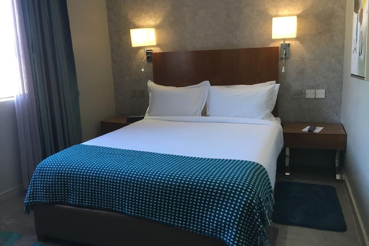 A hotel room with a large bed, two bedside tables, a nightstand, a wall-mounted lamp, and a window with curtains.