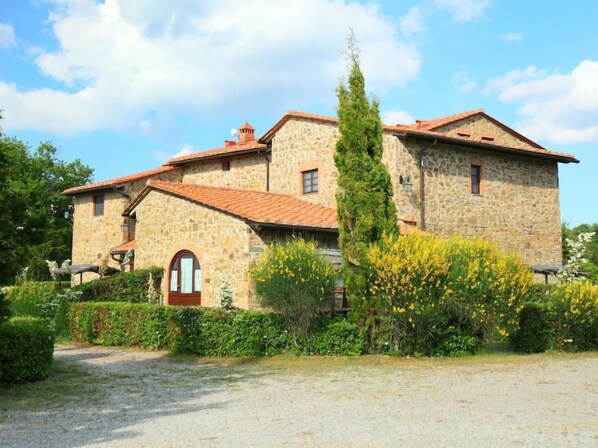 A stone building with a red-tiled roof, a central entrance, and a surrounding garden.