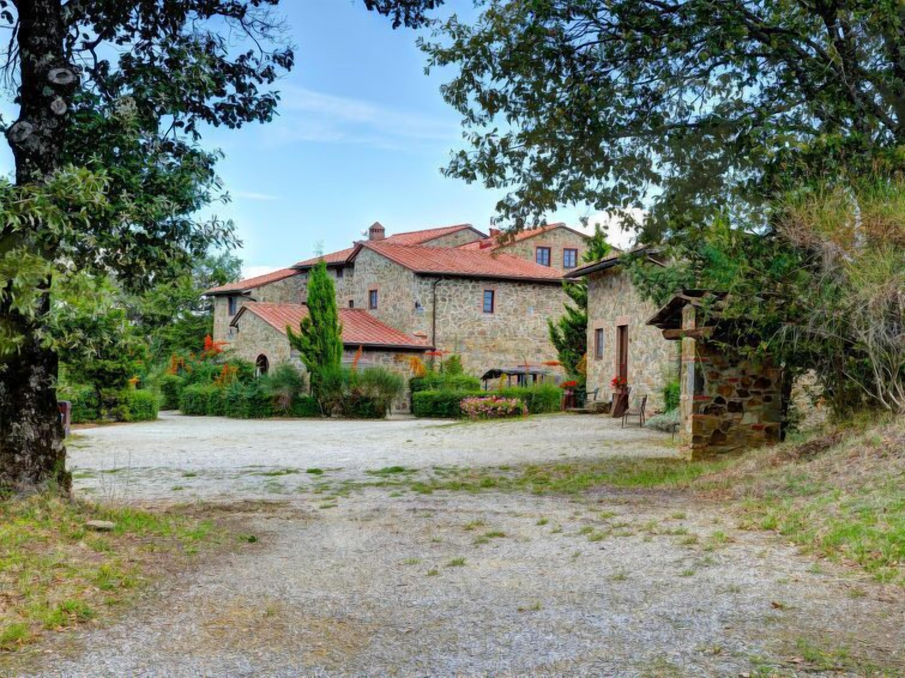 A stone house with a red-tiled roof, surrounded by greenery and a gravel driveway.