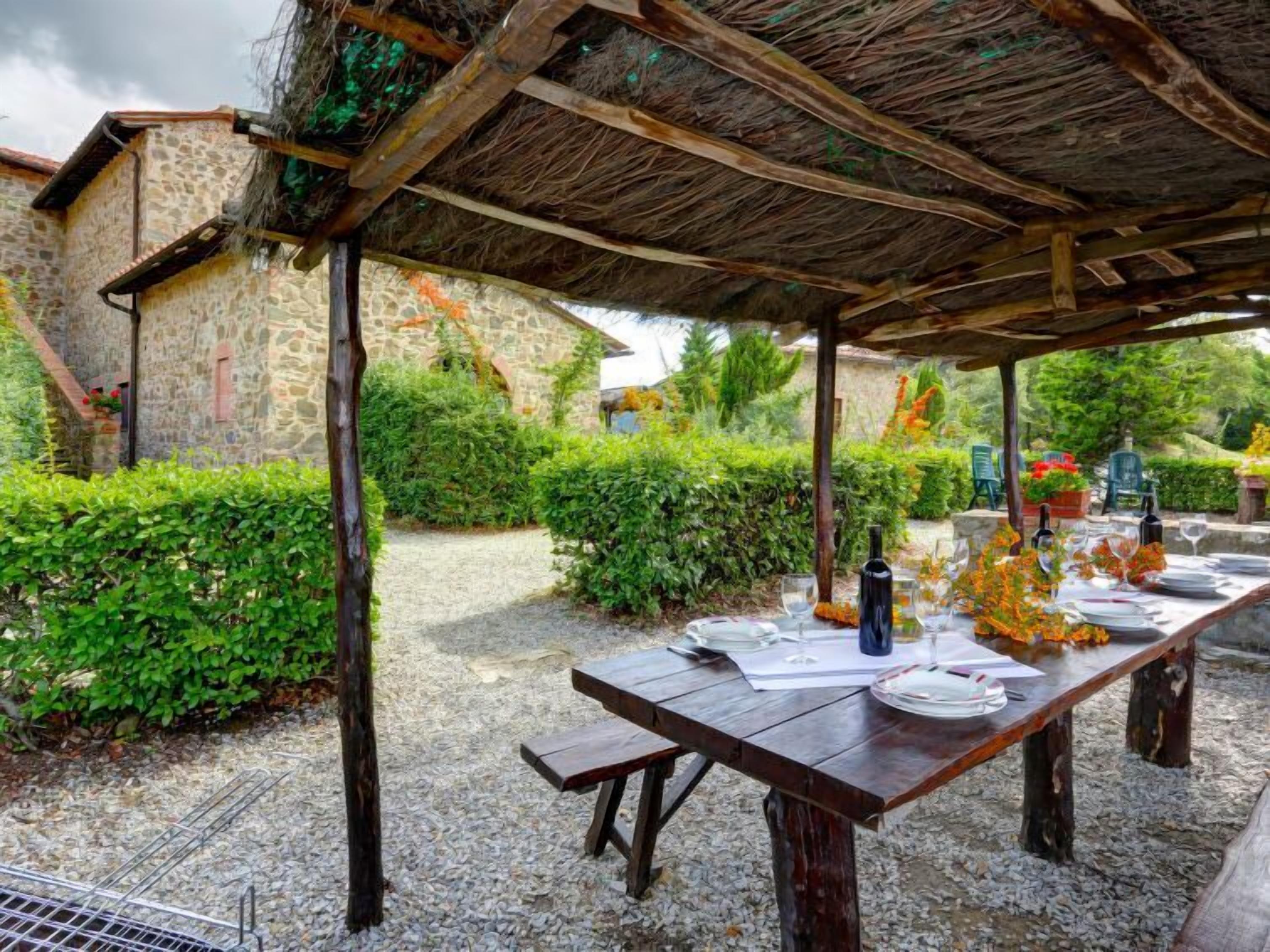 An outdoor dining area with a rustic stone building in the background, a wooden table set for a meal, and a thatched roof overhead.
