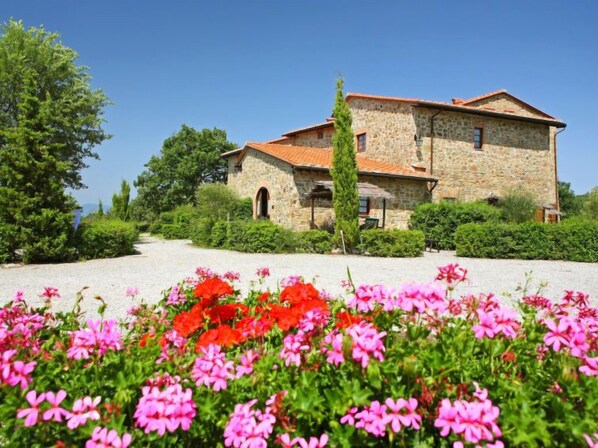 Un bâtiment en pierre, surmonté d’un toit de tuiles rouges, entouré de verdure et de plantes en fleurs.