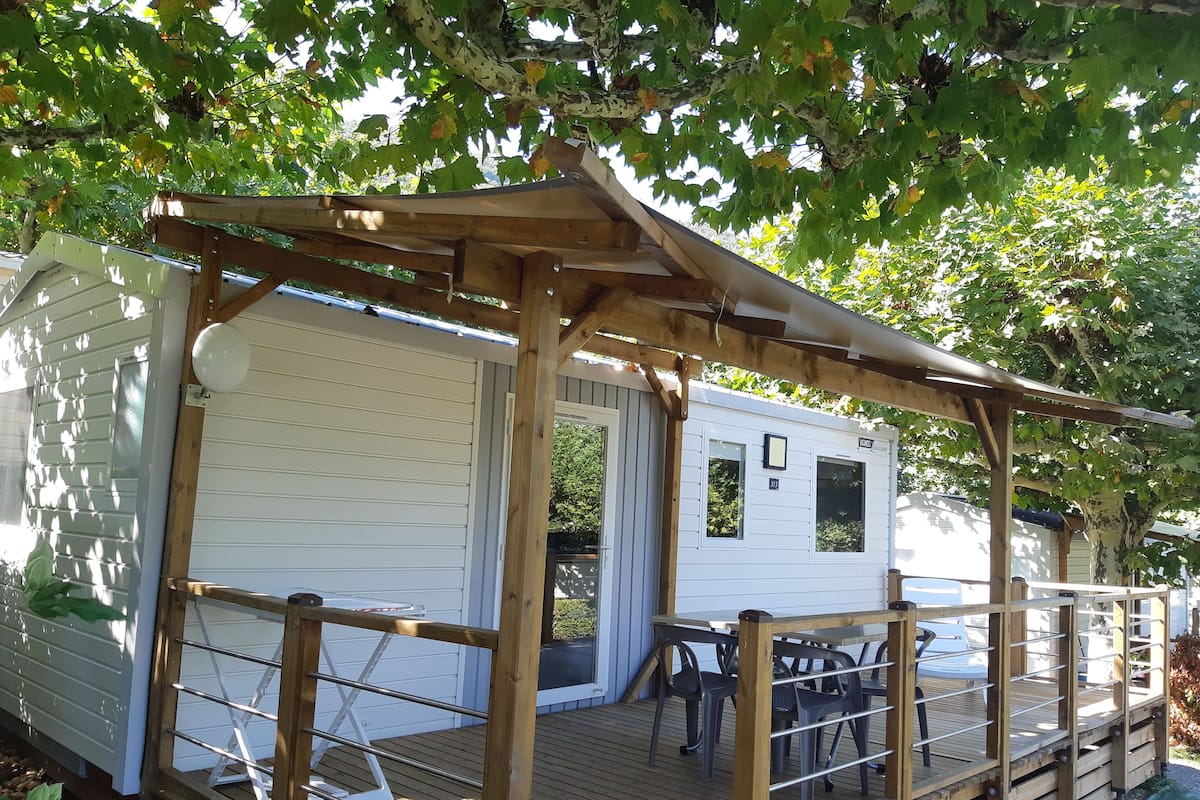 A wooden deck with a pergola, a small white mobile home with a window, and a table with chairs.