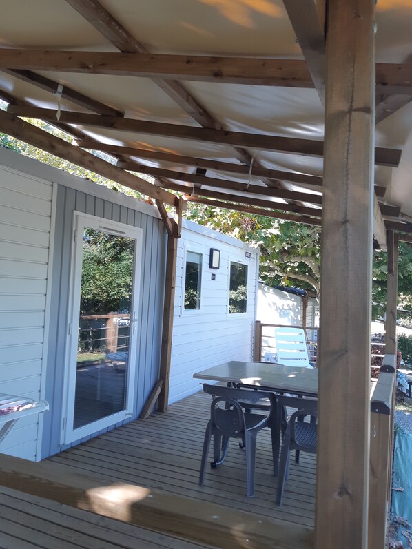 A covered patio area with a wooden deck, a table with chairs, and a white building with a sliding door.