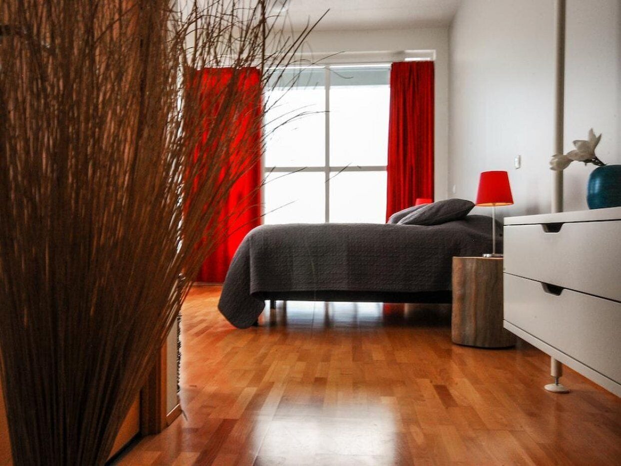 Un dormitorio moderno con piso de madera, una cama con ropa de cama gris, un tocador blanco y una lámpara roja.