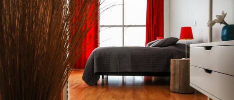 Un dormitorio moderno con piso de madera, una cama con ropa de cama gris, un tocador blanco y una lámpara roja.