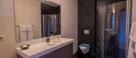 Un baño moderno con un tocador blanco, un espejo grande, cabina de ducha de vidrio y una pared de acento oscura.