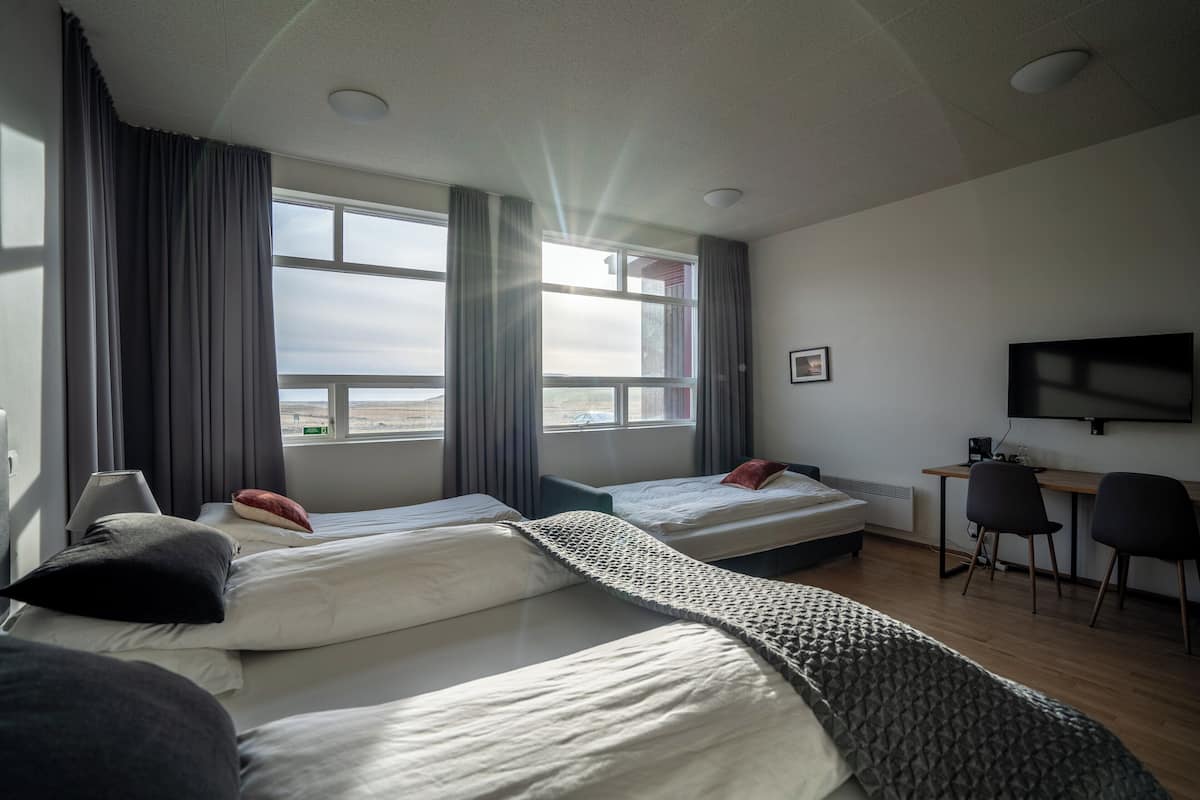 A hotel room with two beds, a desk, a chair, a TV, and large windows with curtains.