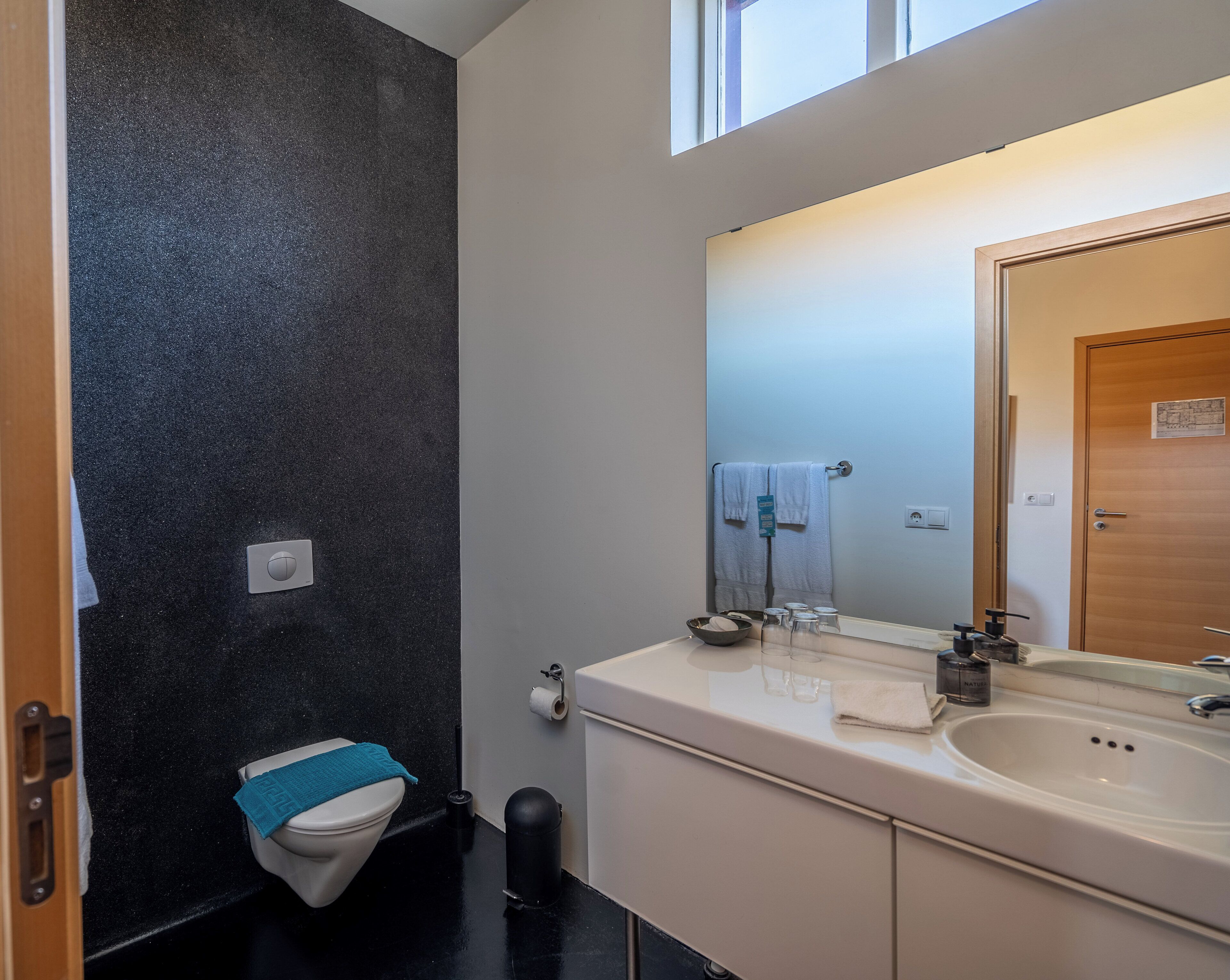 A modern bathroom with a black textured wall, a white sink, a mirror, and a toilet with a blue towel.