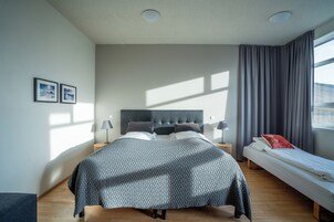 A modern bedroom with a large bed, a smaller bed, a nightstand, a lamp, and a window with curtains.