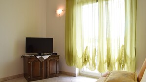 Une pièce avec un téléviseur posé sur un meuble en bois, un lit avec un oreiller jaune et une fenêtre ornée de rideaux légers.