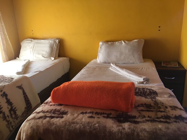 Habitación de hotel con dos camas, cada una con sábanas y almohadas blancas, una manta estampada en la cama del primer plano y una revista sobre la mesita de noche.