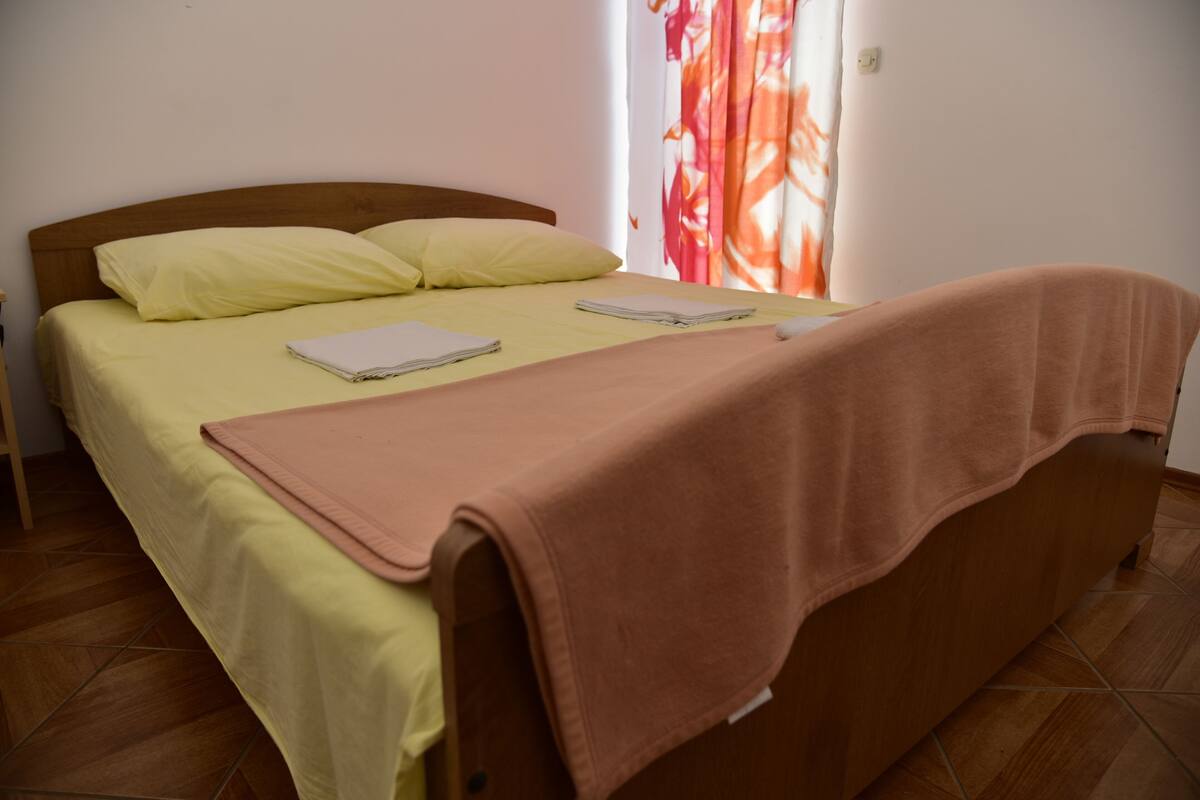 Un lit bien fait, avec une couvre-lit jaune et une couverture marron, une tête de lit en bois et une fenêtre ornée de rideaux rouges.