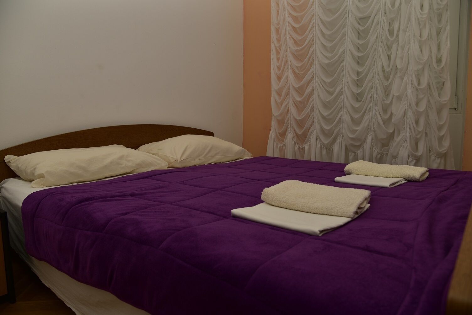 A neatly made bed with a purple bedspread and white pillows, a wooden headboard, and a white curtain with a wavy pattern.