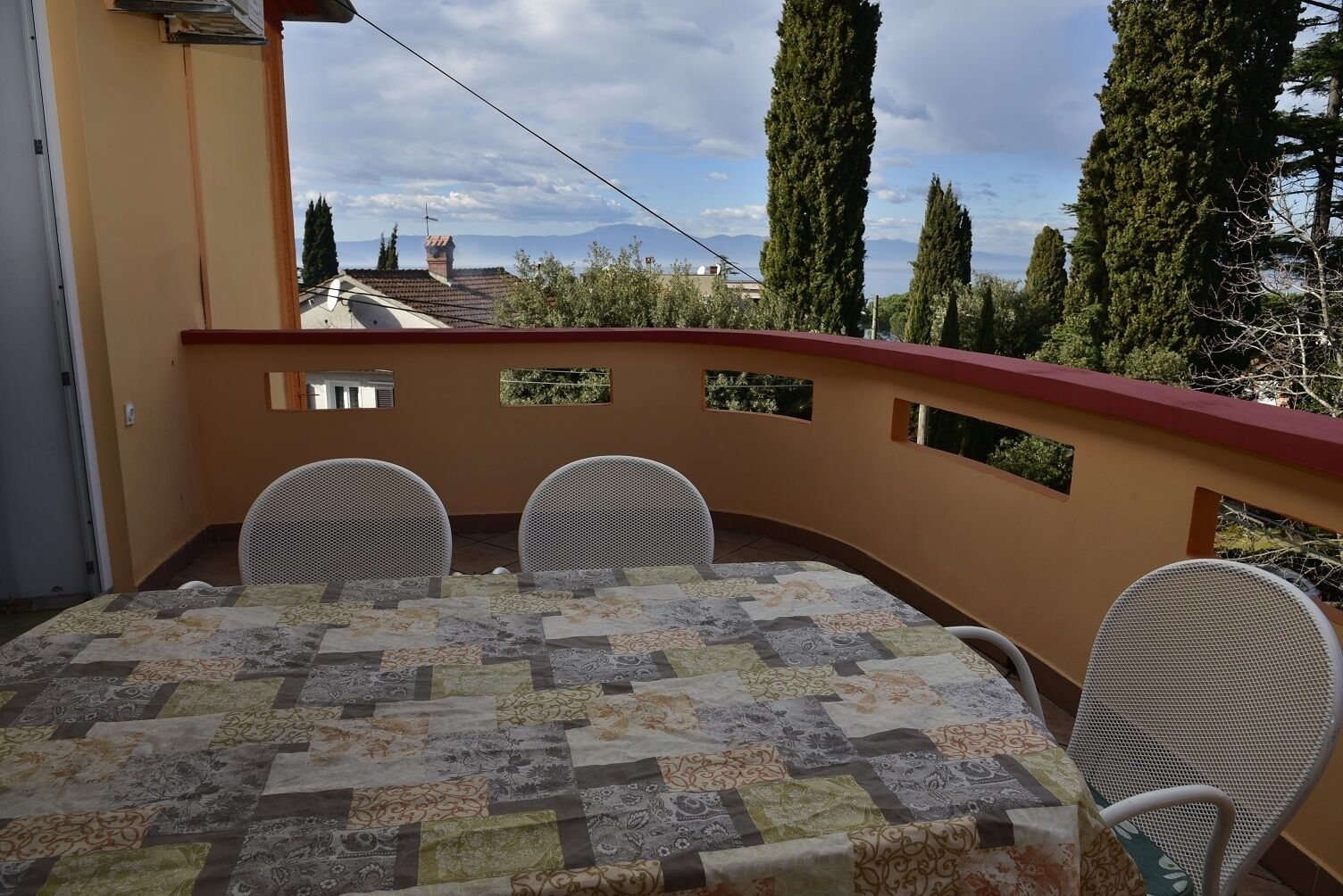 Un balcone con un tavolo ricoperto da una tovaglia patchwork, tre sedie in vimini e una ringhiera rossa.