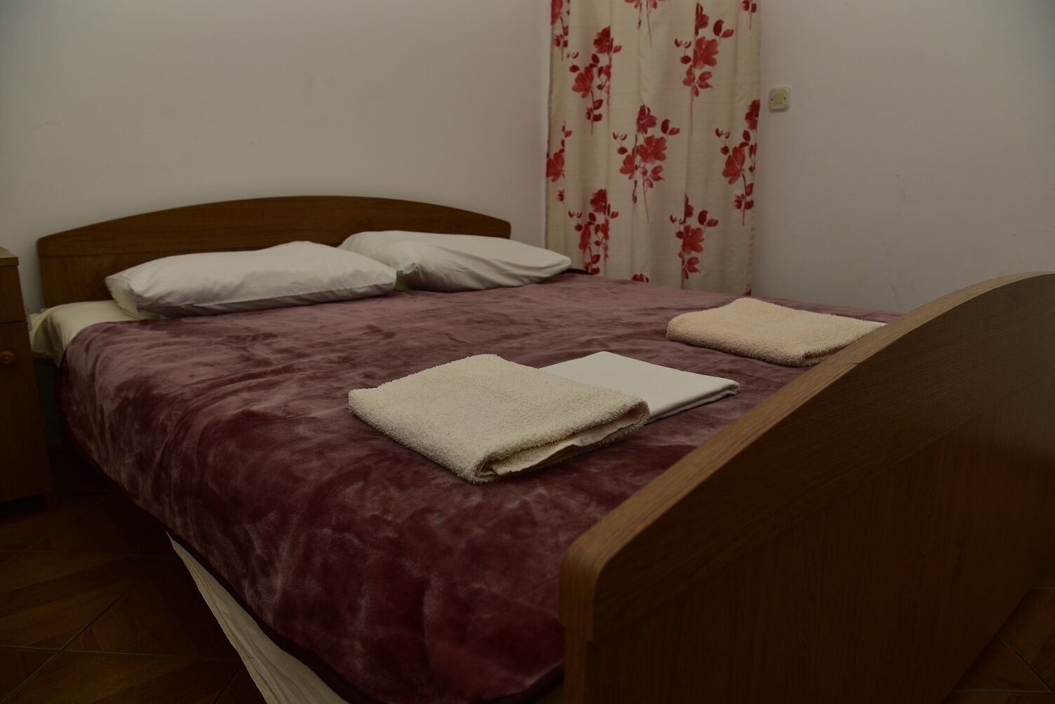 Uma cama de solteiro com cabeceira de madeira, um cobreleite vermelho, duas almofadas e duas toalhas dobradas.