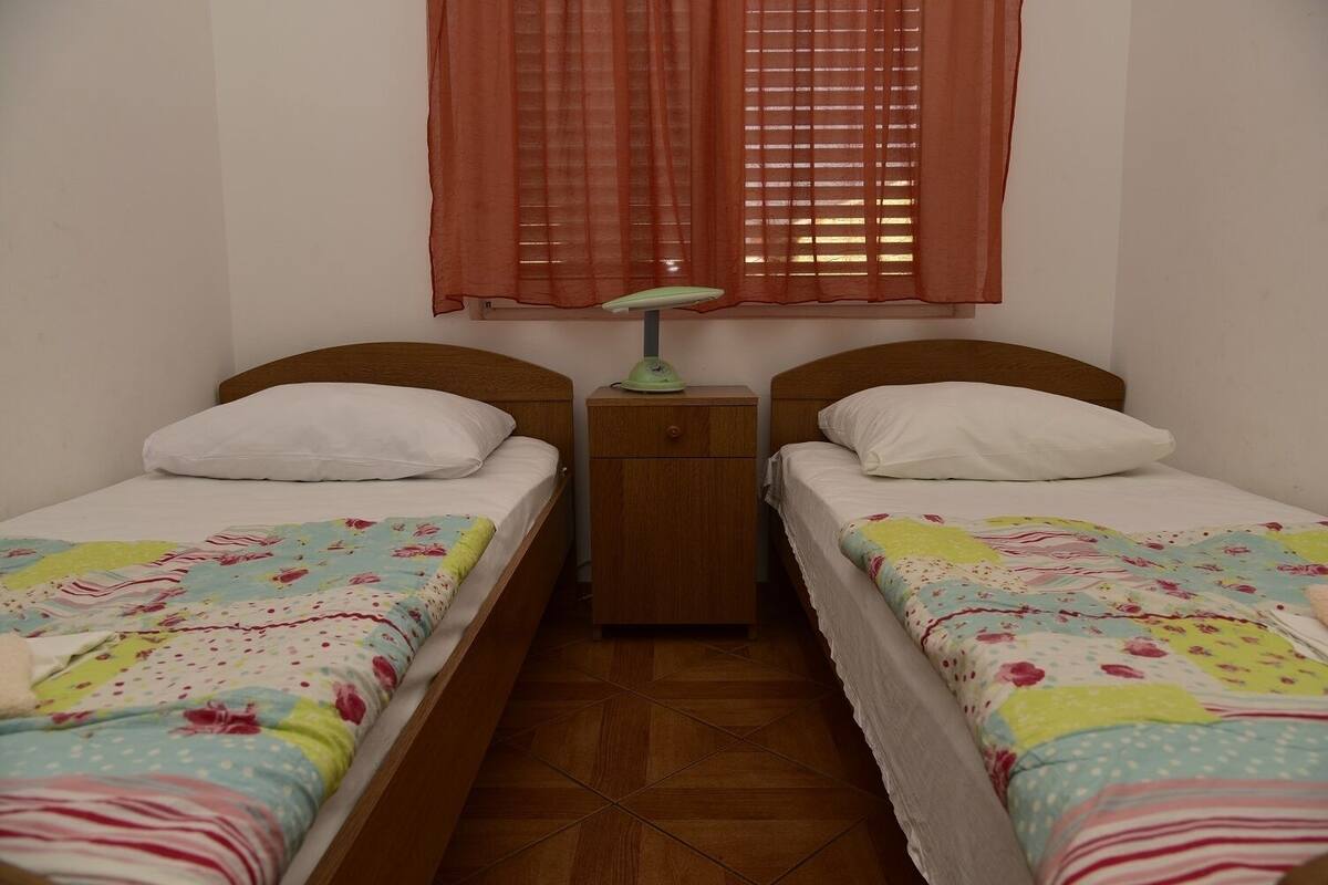 Une petite chambre propre, avec deux lits simples, une table de chevet, une lampe et une fenêtre avec des rideaux.
