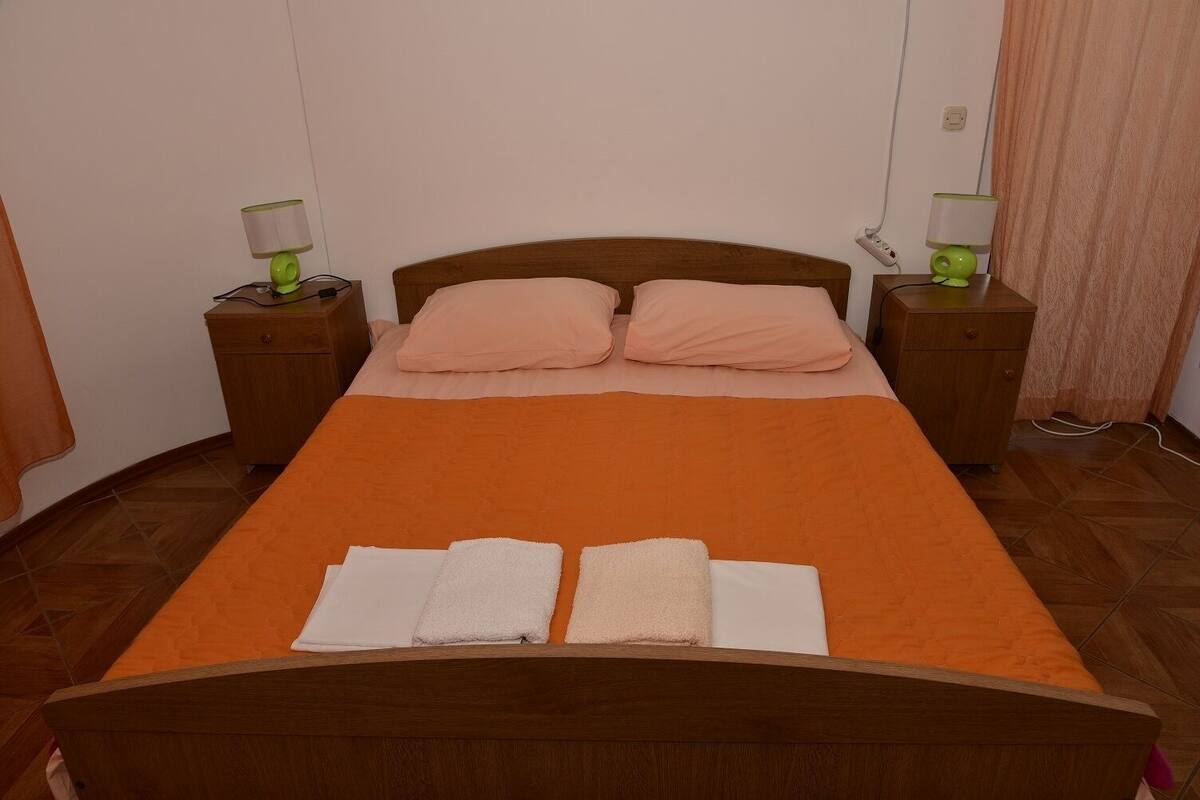Un lit bien fait, recouvert d’une couverture orange, avec deux oreillers et trois serviettes pliées. Il y a deux tables de chevet avec des lampes, et une prise électrique sur le mur.